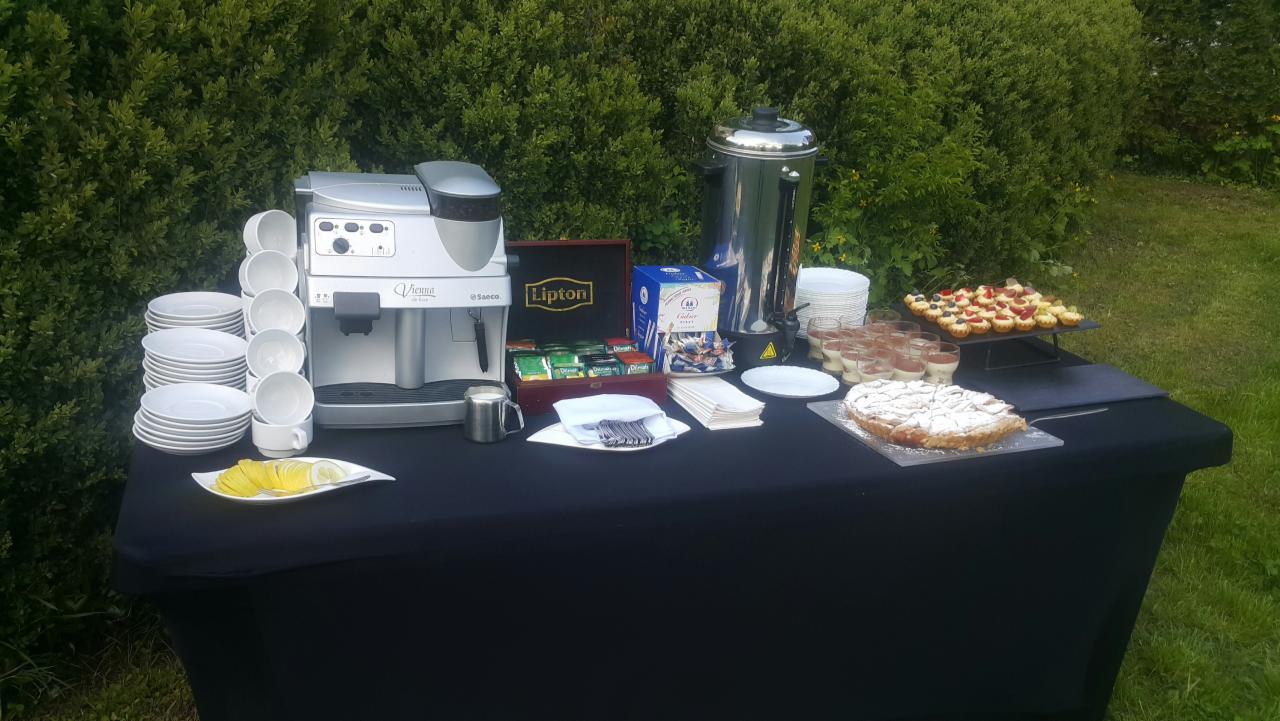 Stół cateringowy z ekspresem do kawy, termosem, deserami i herbatą Lipton na tle zieleni.