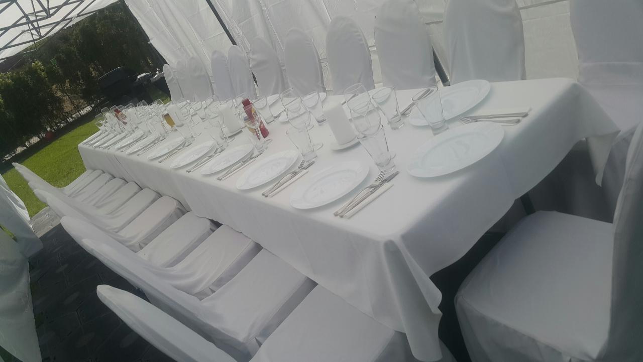 Długi stół nakryty białym obrusem, z równo ułożonymi talerzami, sztućcami i szklankami, otoczony krzesłami w białych pokrowcach, ustawiony w namiocie z widokiem na zieleń.