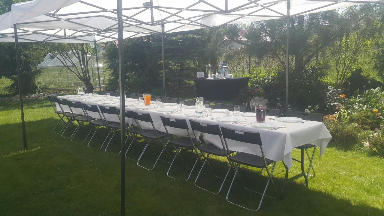 Długi stół nakryty białym obrusem i zastawą dla wielu osób, ustawiony na trawie pod białym namiotem, w tle ogród z zielenią i zabudowaniami.