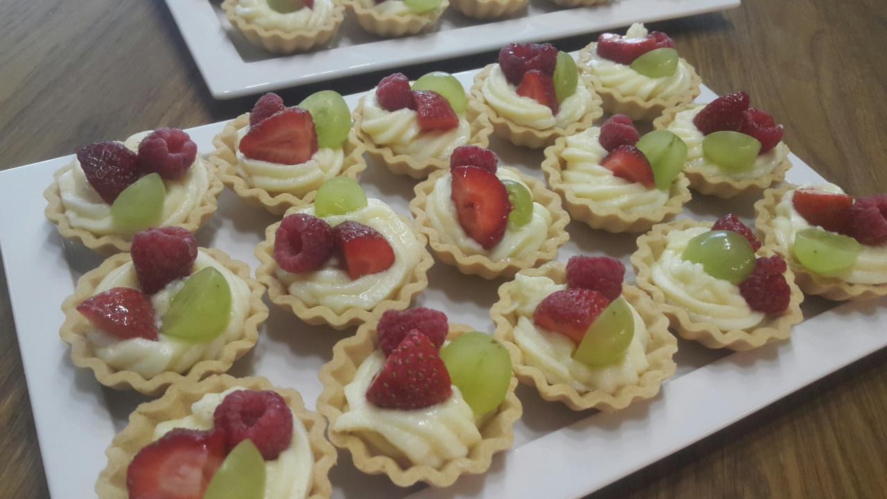 Tarty z kremem i owocami: malinami, truskawkami i zielonymi winogronami, ułożone na białym prostokątnym talerzu na drewnianym stole.