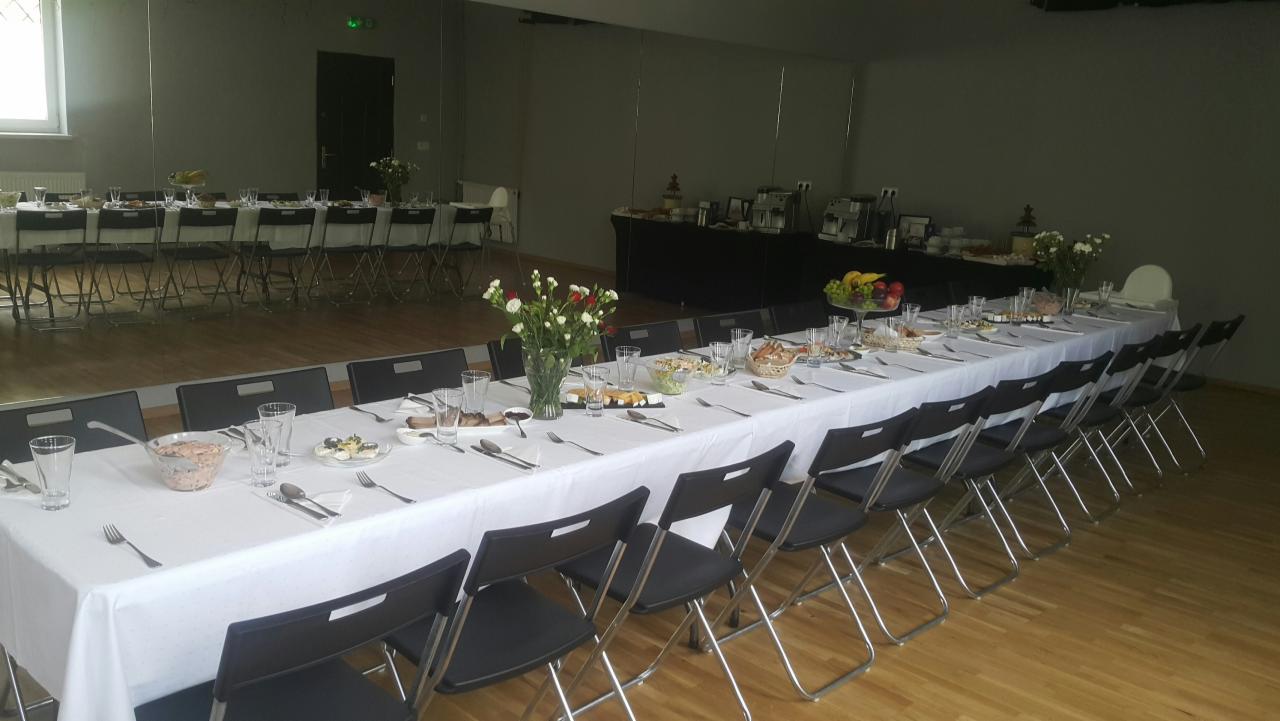 Długi stół nakryty białym obrusem z ustawionymi szklankami, sztućcami i różnorodnymi potrawami, otoczony czarnymi krzesłami w przestronnym pomieszczeniu z parkietem i lustrzaną ścianą.