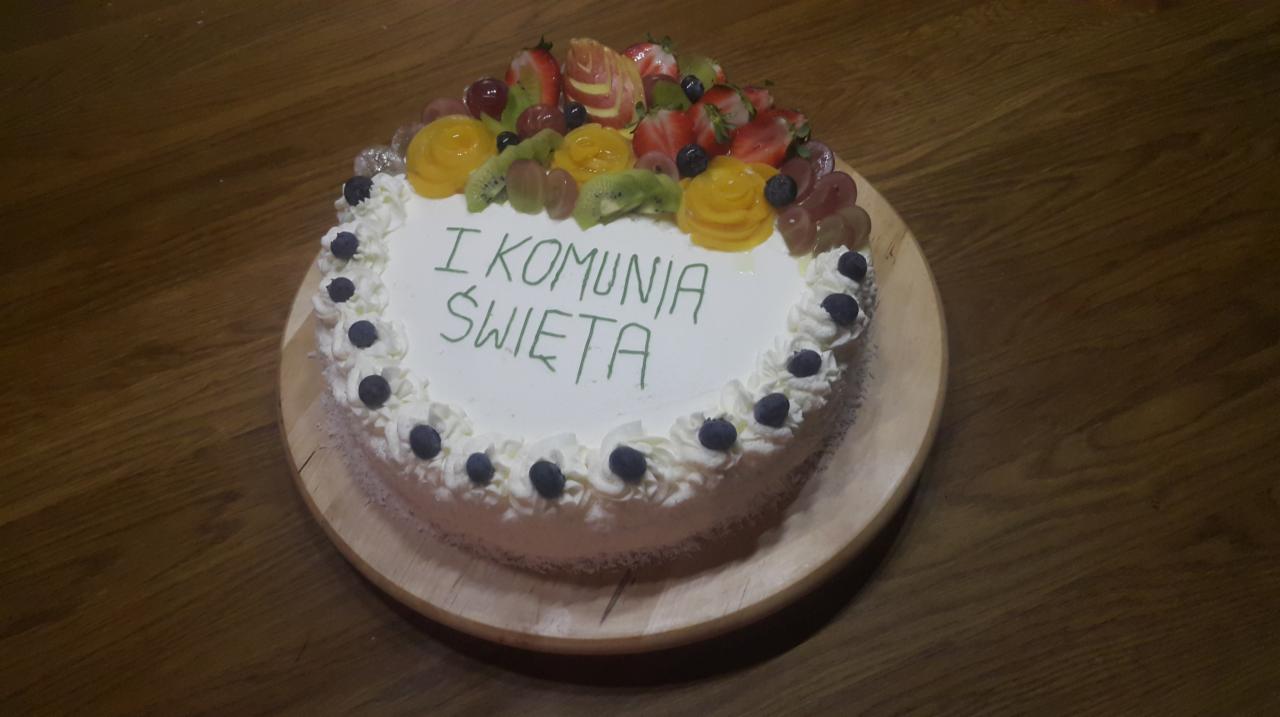 Tort na komunię z owocami: truskawki, winogrona, kiwi, brzoskwinie, udekorowany kremem i borówkami, leżący na drewnianej desce na drewnianym stole.