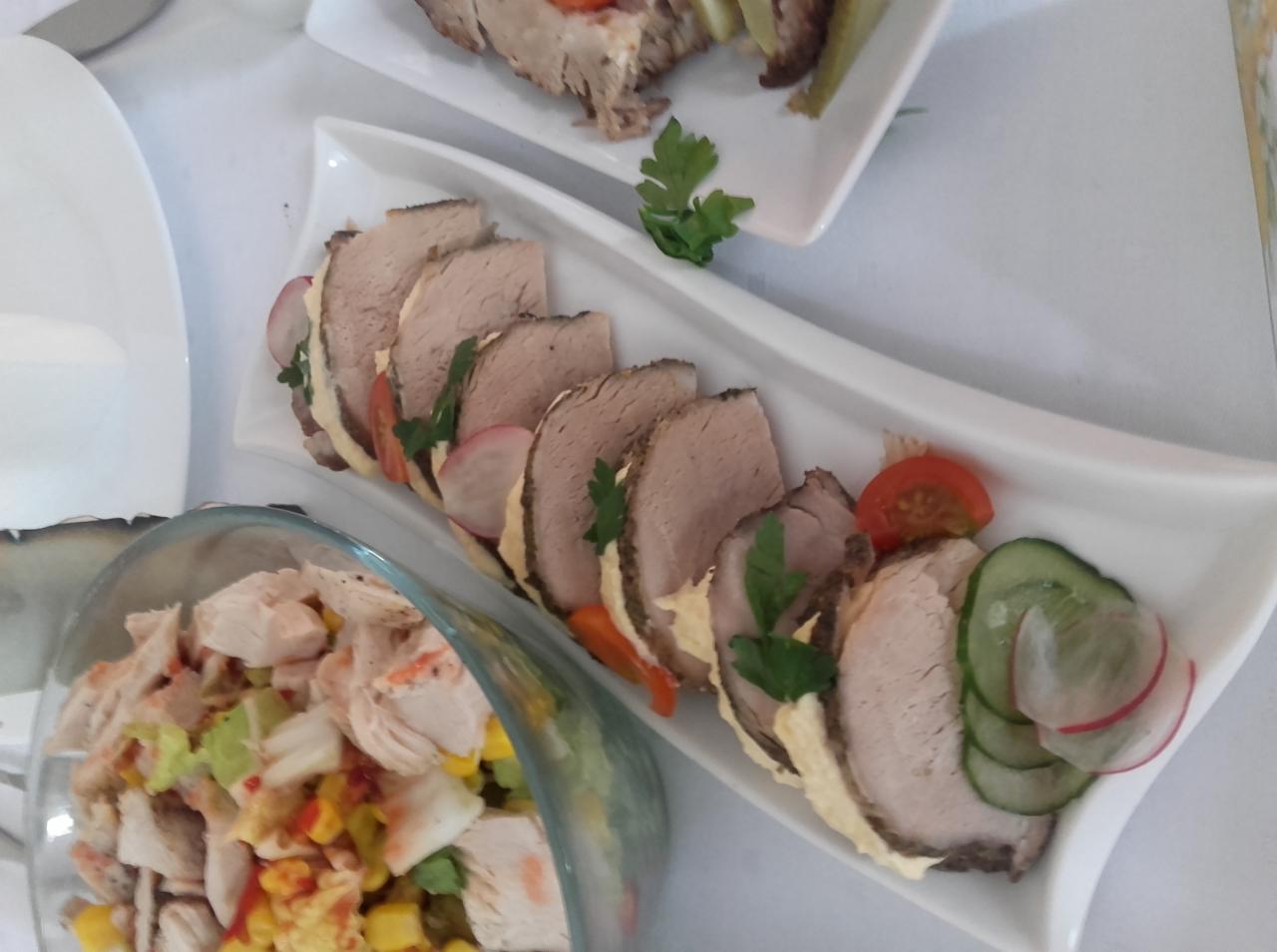 Elegancko ułożone plastry pieczonego mięsa z kremowym sosem, udekorowane rzodkiewką, ogórkiem i pomidorem na białym, falistym talerzu, obok szklana misa z sałatką z kurczakiem, kukurydzą i papryką.