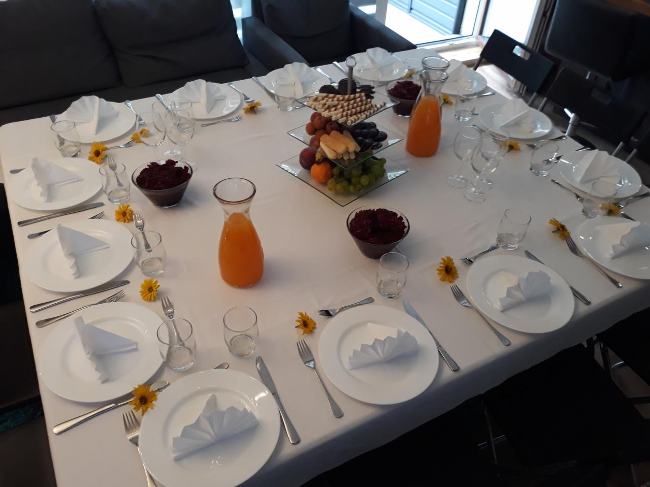 Nakryty stół z białym obrusem, zastawą, szklankami, sztućcami, dzbankiem z pomarańczowym napojem, miseczkami z buraczkami i paterą z owocami i słodyczami, udekorowany żółtymi kwiatami.