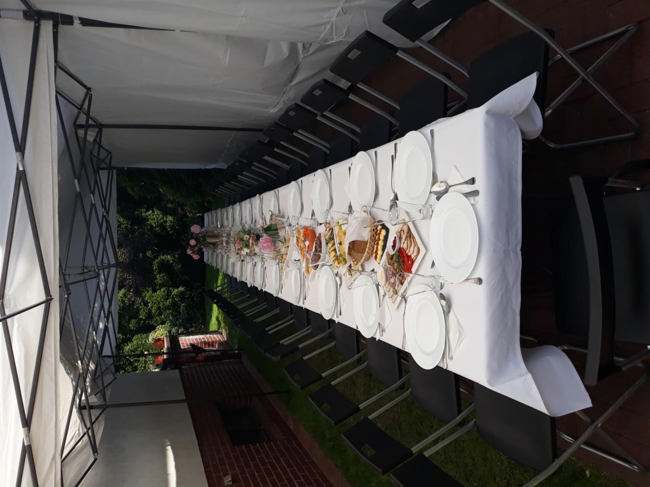 Długi stół pod namiotem, nakryty białym obrusem, z ustawionymi talerzami, sztućcami, przekąskami i dekoracjami kwiatowymi, przygotowany na przyjęcie na zewnątrz.