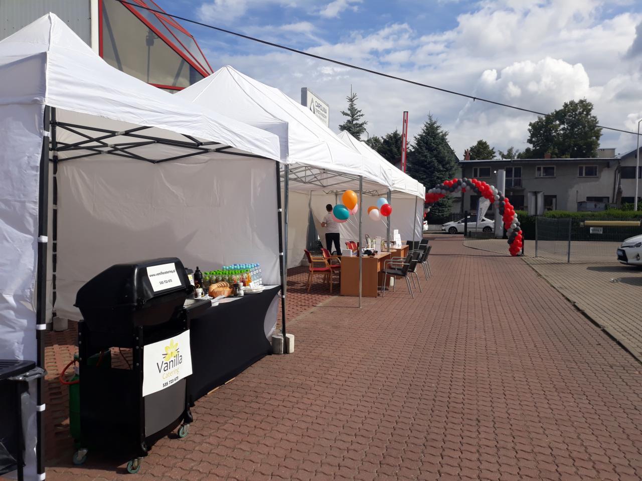 Stanowisko cateringowe z grillem i napojami przed białymi namiotami, udekorowane balonami, z widocznym wejściem na teren firmy, na tle błękitnego nieba.