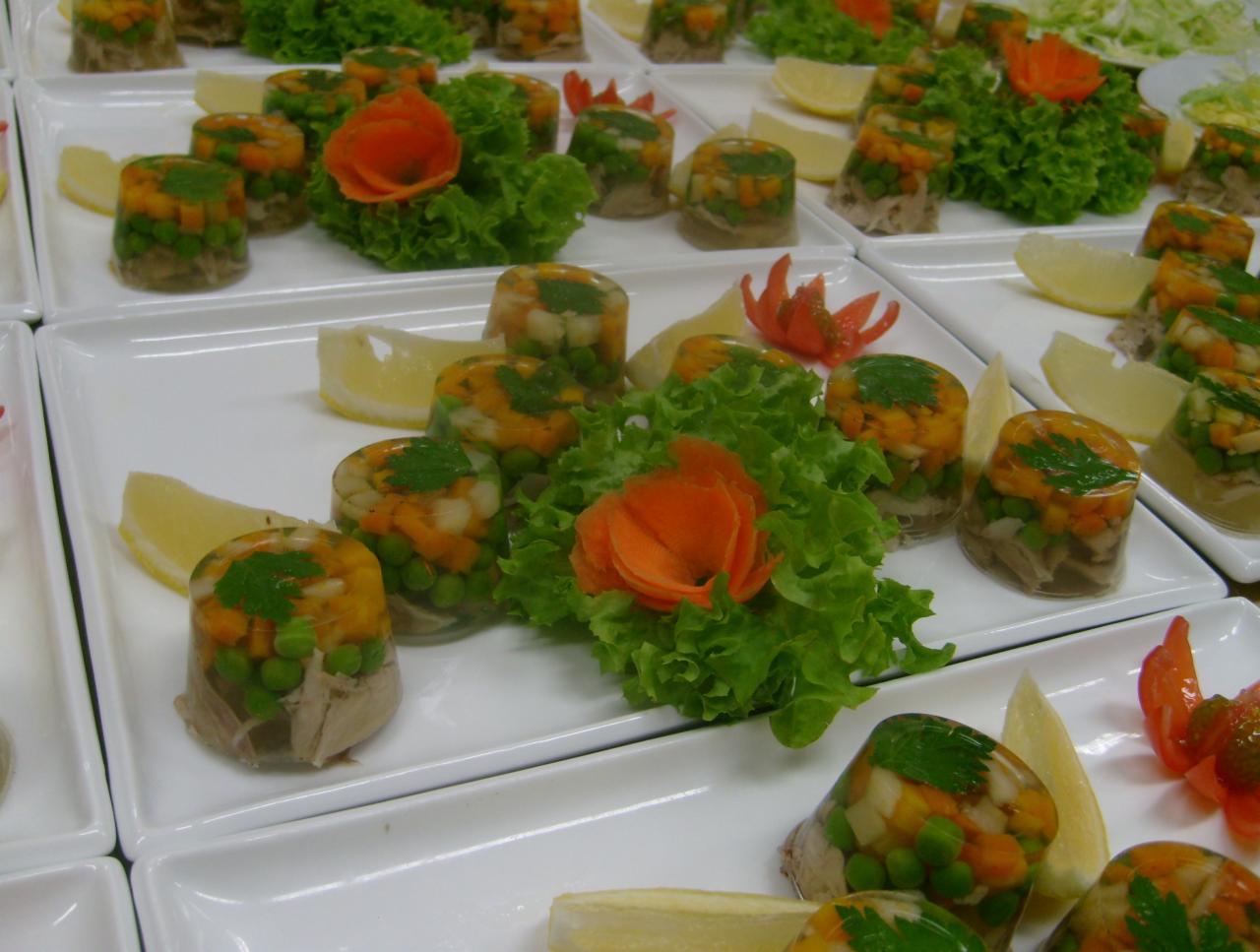 Przystawki w galarecie ułożone na białych talerzach, dekorowane sałatą i plasterkami cytryny, z akcentem w postaci rzeźbionych warzyw.