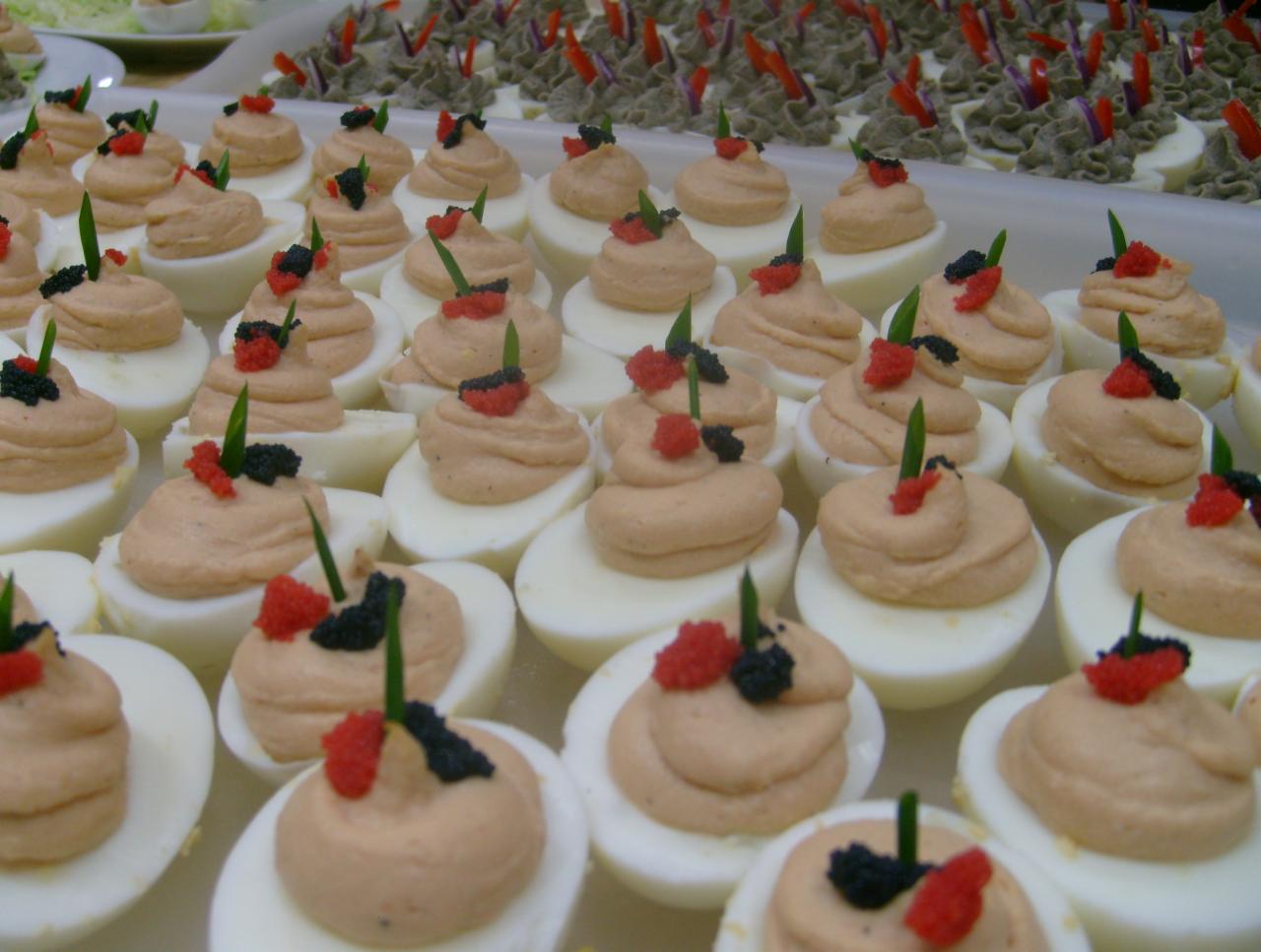 Taca z jajkami na twardo przekrojonymi na pół, udekorowanymi kremowym farszem, czerwoną i czarną posypką oraz szczypiorkiem, ułożonymi w rzędach obok siebie.