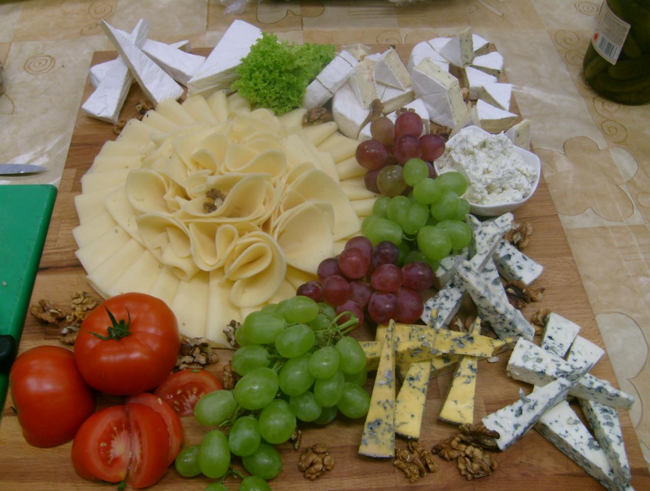 Deska serów z różnymi rodzajami sera, winogronami, pomidorami i orzechami włoskimi, ułożona na drewnianej desce do krojenia.