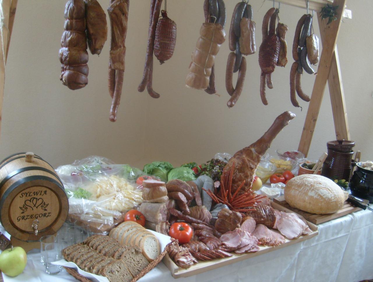 Bogato zastawiony stół z wędlinami, pieczywem, warzywami i dekoracyjnym drewnianym barełkiem z napisem Sylwia i Grzegorz, w tle wiszące wędzonki na drewnianym stelażu.