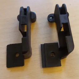 Dwa czarne, plastikowe elementy z widocznymi śladami druku 3D, leżące na jasnym, drewnianym blacie. Jeden element ma wytłoczoną literę 'R'.