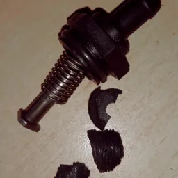 Uszkodzony, rozłożony zawór zwrotny z metalową sprężyną i fragmentami gumowej membrany leżący na beżowym tle.