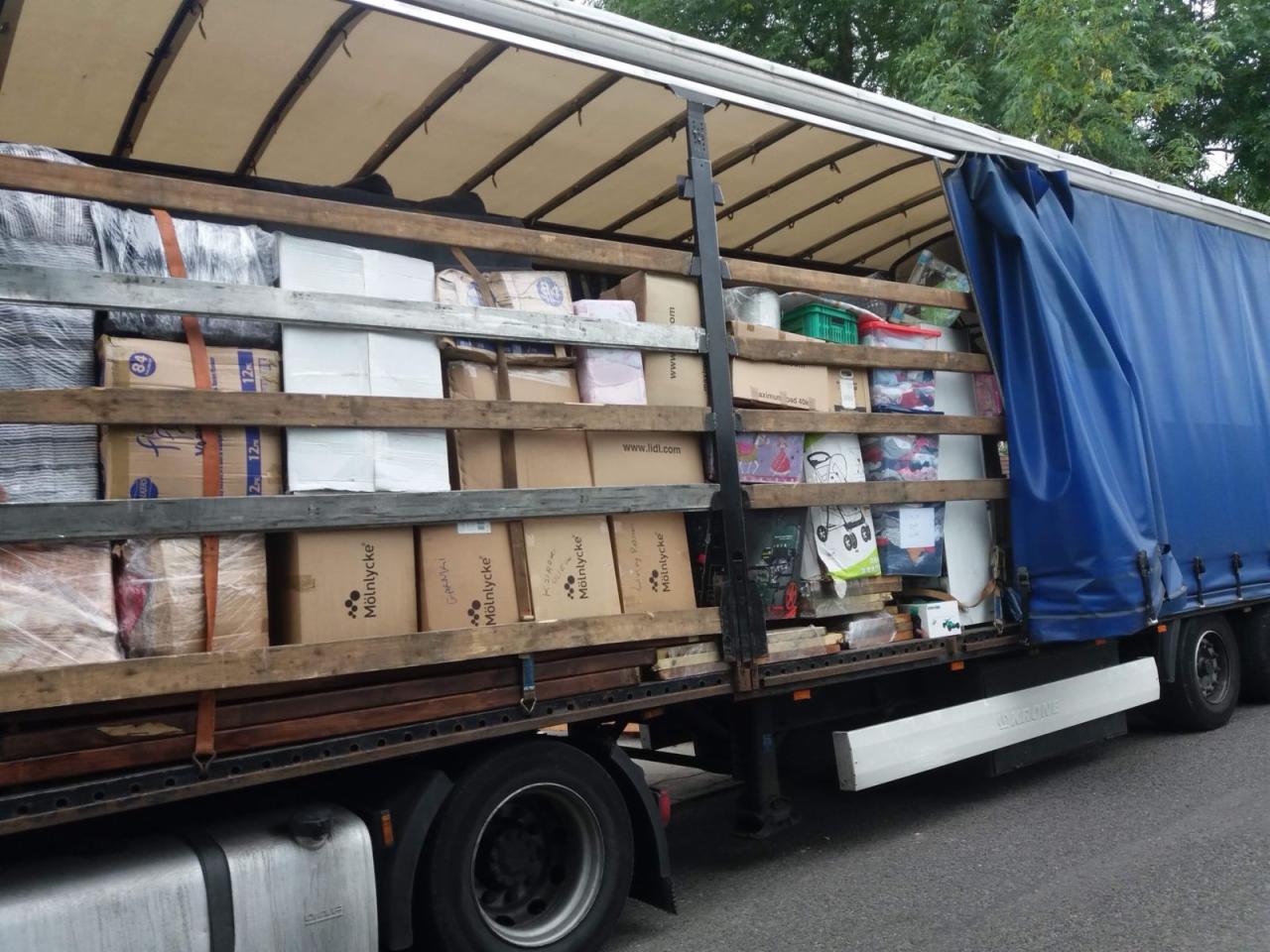 Ciężarówka z otwartą naczepą załadowana różnorodnymi kartonami i pakunkami, zabezpieczona drewnianymi poprzeczkami, widok z boku na tle zieleni.