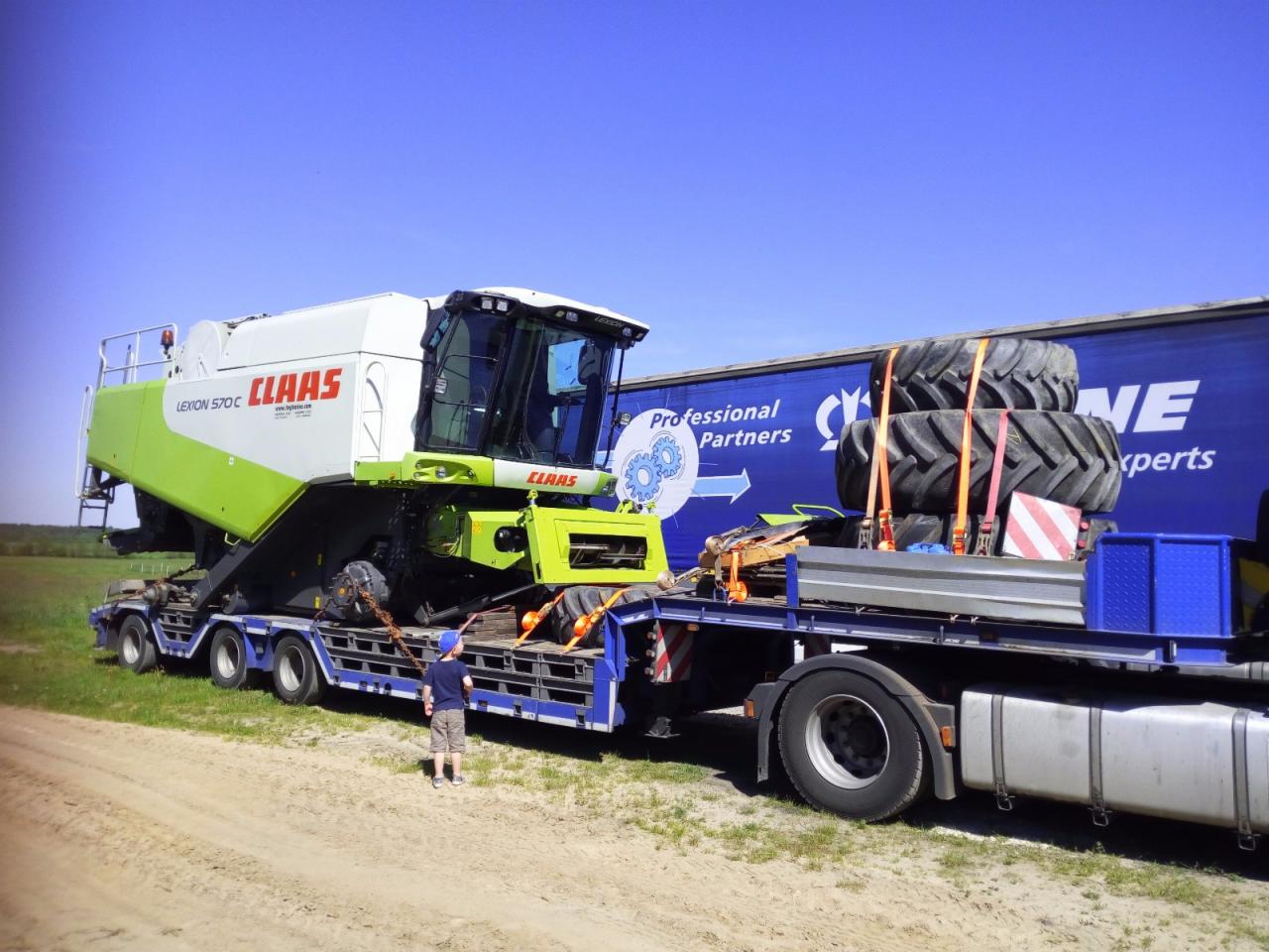 Transport niskopodwoziowy: Kombajn rolniczy Claas Lexion 570 C zabezpieczony pasami na platformie, obok stoi dziecko w niebieskiej czapce, na naczepie dodatkowe opony.