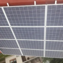 Panele fotowoltaiczne zamontowane na czerwonym dachu, widok z góry, w tle budynek mieszkalny.