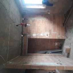 Wnętrze w trakcie prac wykończeniowych: widoczne przewody elektryczne wystające ze ściany z cegieł, tymczasowe oświetlenie budowlane, surowa betonowa ściana po lewej stronie oraz częściowo ułożona...