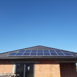 elektroinstalm - Niedokończony dom z cegły z zamontowanymi panelami słonecznymi na dachu pokrytym czarną dachówką, drabina oparta o dach, błękitne niebo w tle.