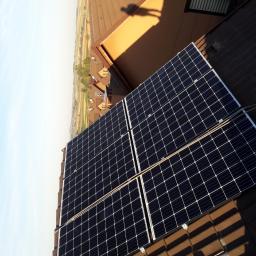 elektroinstalm - Cztery panele słoneczne zamontowane na brązowym dachu z widokiem na okoliczne domy i pola w oddali, cień osoby widoczny na sąsiednim dachu.