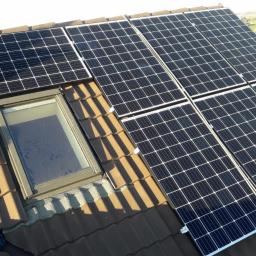 elektroinstalm - Panele słoneczne zamontowane na brązowym dachu z dachówką, obok okna dachowego; widok z góry, z widocznymi elementami mocującymi i fragmentem niebieskiej tkaniny w rogu.