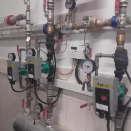 Kompleksowa instalacja grzewcza z licznymi rurami, zaworami, manometrami i pompami obiegowymi, zamontowana na ścianie z białymi kafelkami.