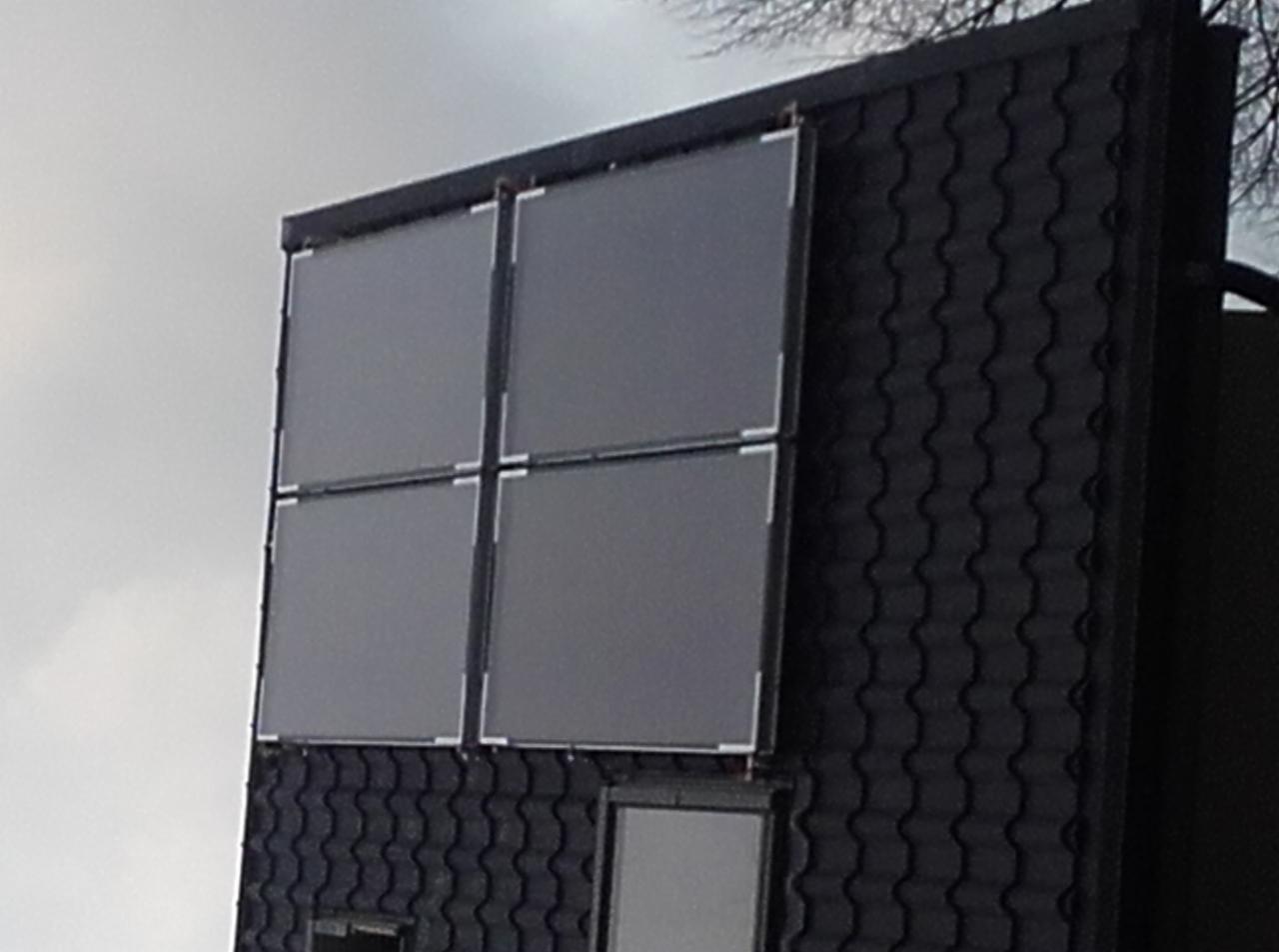 Cztery panele słoneczne w metalowych ramach zamontowane na ciemnym, skośnym dachu pokrytym dachówką bitumiczną, widoczne z dołu pod kątem, przy pochmurnym niebie.