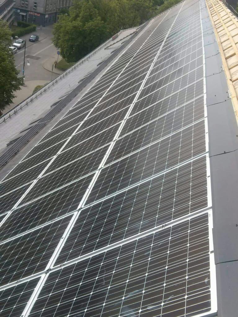 Rozległy dach pokryty panelami słonecznymi, widok z góry, z fragmentem ulicy i budynków w tle.
