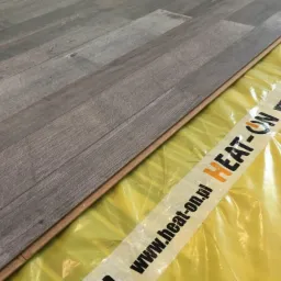 Panel podłogowy o strukturze drewna układany na żółtej folii z napisem 'HEAT-ON'