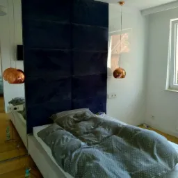 Nowoczesna sypialnia z łóżkiem z wezgłowiem z ciemnoniebieskich paneli tapicerowanych, wiszącymi lampami w kolorze miedzi i lustrem na ścianie.