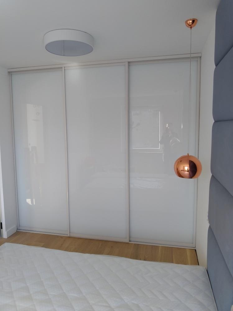 Biała szafa przesuwna z trzema drzwiami, widoczna z boku łóżka z pikowanym zagłówkiem i miedzianą lampą wiszącą.