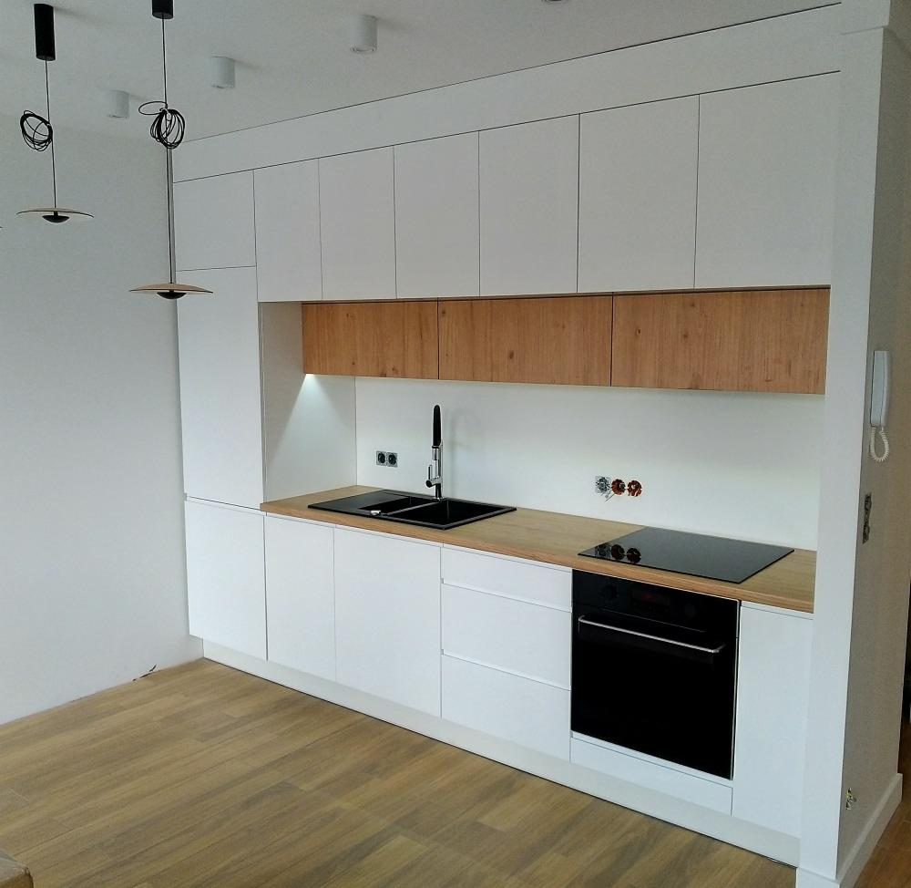 Nowoczesna kuchnia na wymiar z białymi frontami szafek i drewnianymi górnymi szafkami, czarny zlew i płyta indukcyjna, minimalistyczne lampy wiszące.