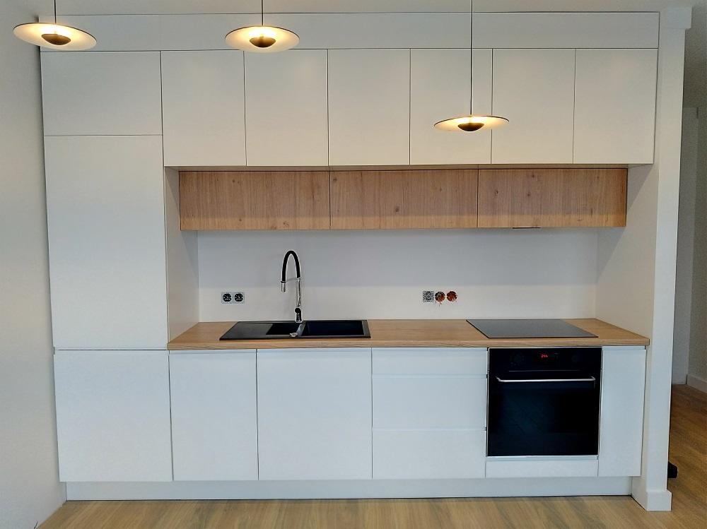 Nowoczesna kuchnia z białymi szafkami i drewnianymi akcentami, oświetlona trzema okrągłymi lampami sufitowymi, z czarnym zlewem i płytą indukcyjną.