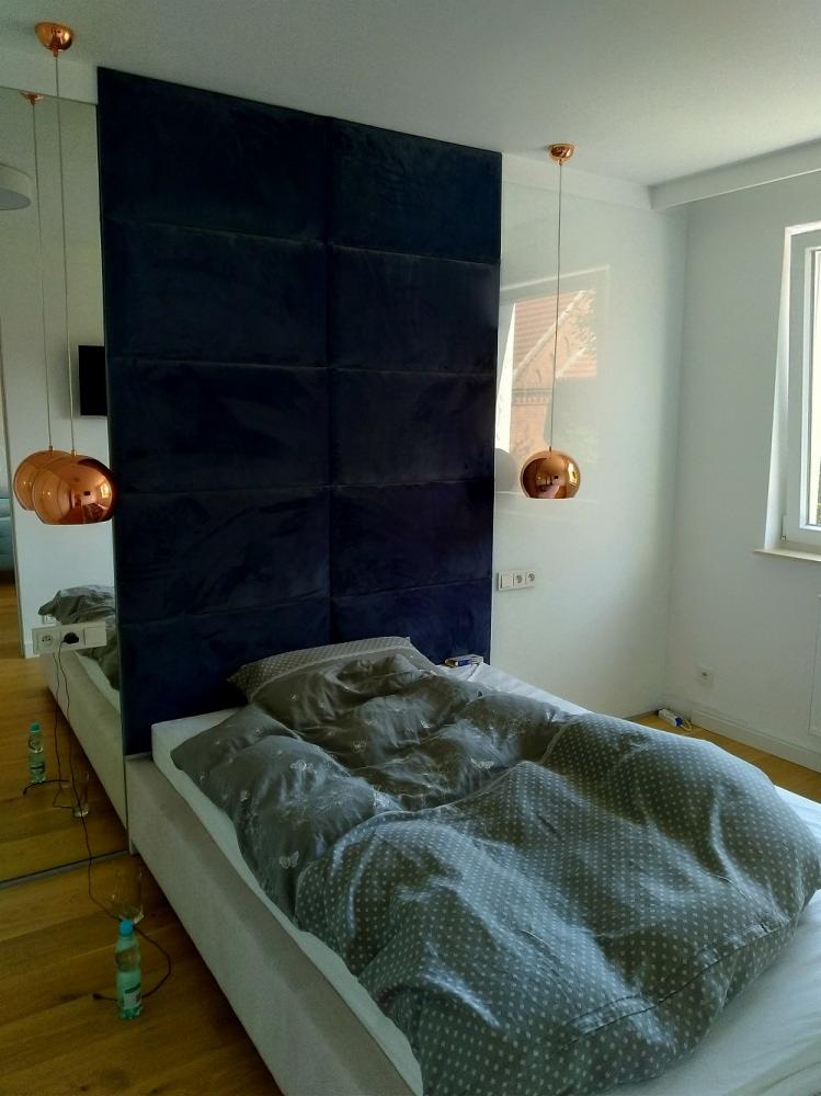 Nowoczesna sypialnia z łóżkiem z wezgłowiem z ciemnoniebieskich paneli tapicerowanych, wiszącymi lampami w kolorze miedzi i lustrem na ścianie.