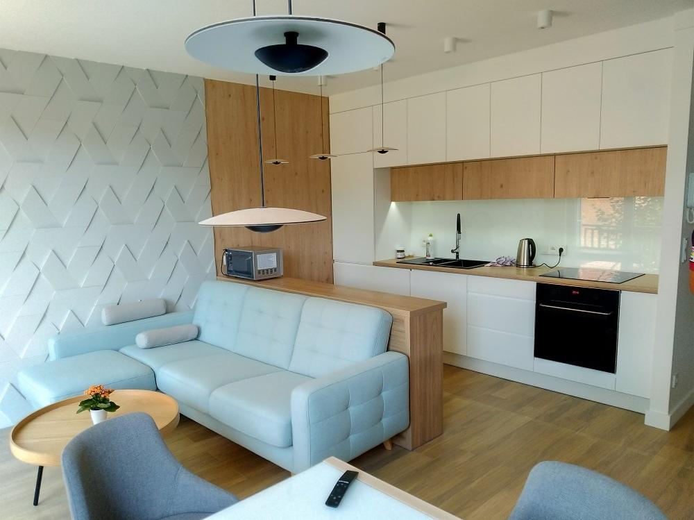 Jasna, nowoczesna kuchnia z drewnianymi akcentami i strefą wypoczynkową: błękitny narożnik z mikrofali umieszczoną nad nim, wiszące lampy dyskretnie oświetlają blat.