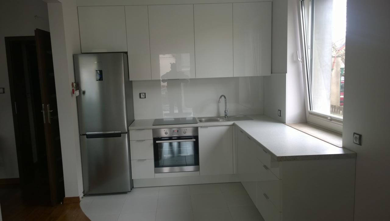 Jasna kuchnia z białymi, błyszczącymi szafkami, srebrną lodówką i blatem przy oknie, widok na płytę grzewczą i zlewozmywak.