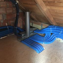 Adrian Zając ADBASS - Niebieskie rury wentylacyjne ułożone równolegle i przymocowane metalowymi opaskami w nowo wykończonym poddaszu z ceglaną ścianą i drewnianą konstrukcją dachu.