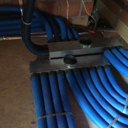 Adrian Zając ADBASS - Rozdzielacz powietrza z licznymi niebieskimi przewodami wentylacyjnymi w surowym otoczeniu poddasza, widoczne elementy konstrukcji drewnianej i ceglanej, przewody spięte metalowymi obejmami.