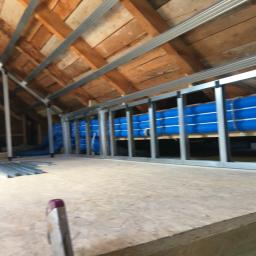 Adrian Zając ADBASS - Poddasze z widoczną konstrukcją metalową i niebieskimi przewodami wentylacyjnymi, częściowo zamontowanymi pod drewnianym stropem, z perspektywy podłogi.