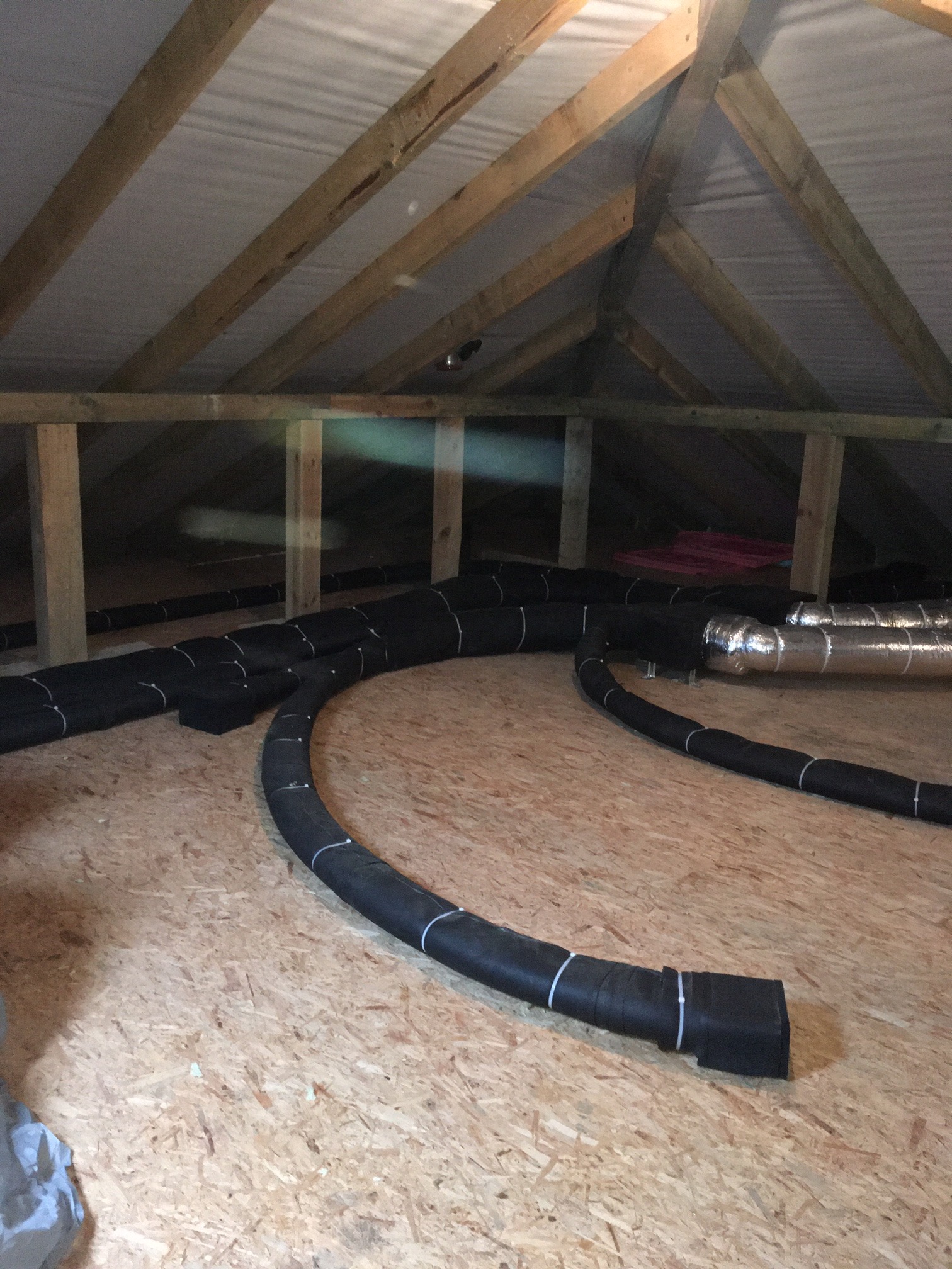 Czarny, izolowany system kanałów wentylacyjnych ułożony na drewnianej podłodze poddasza z widoczną konstrukcją dachu i izolacją.