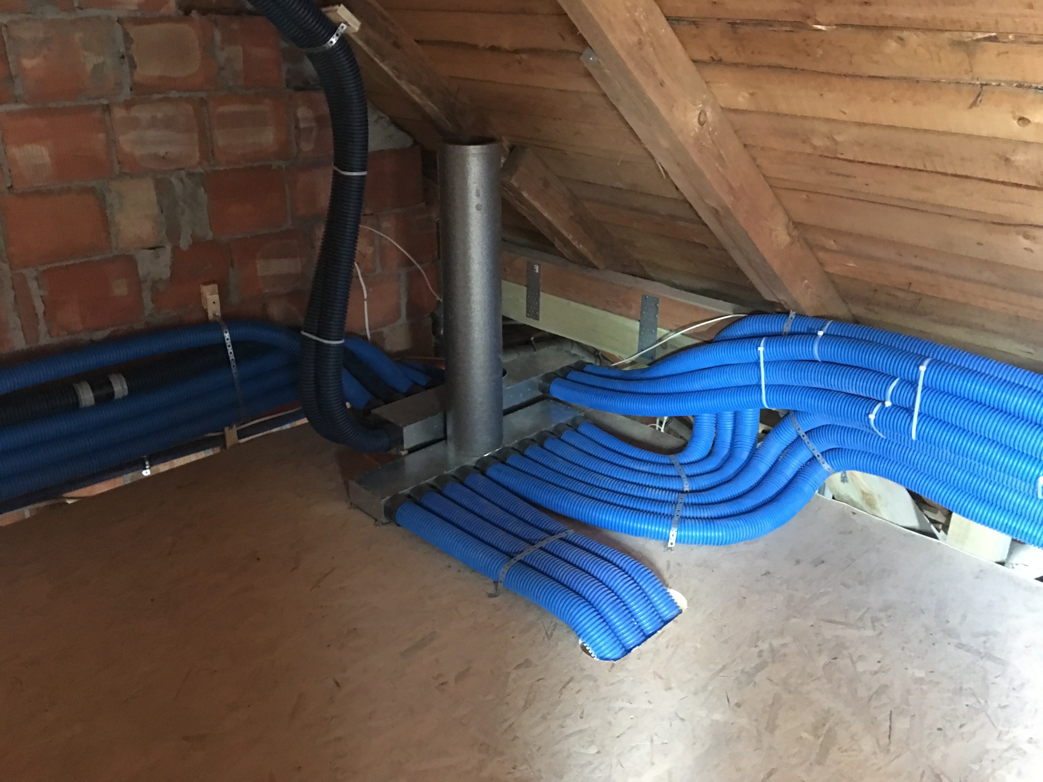 Niebieskie rury wentylacyjne ułożone równolegle i przymocowane metalowymi opaskami w nowo wykończonym poddaszu z ceglaną ścianą i drewnianą konstrukcją dachu.