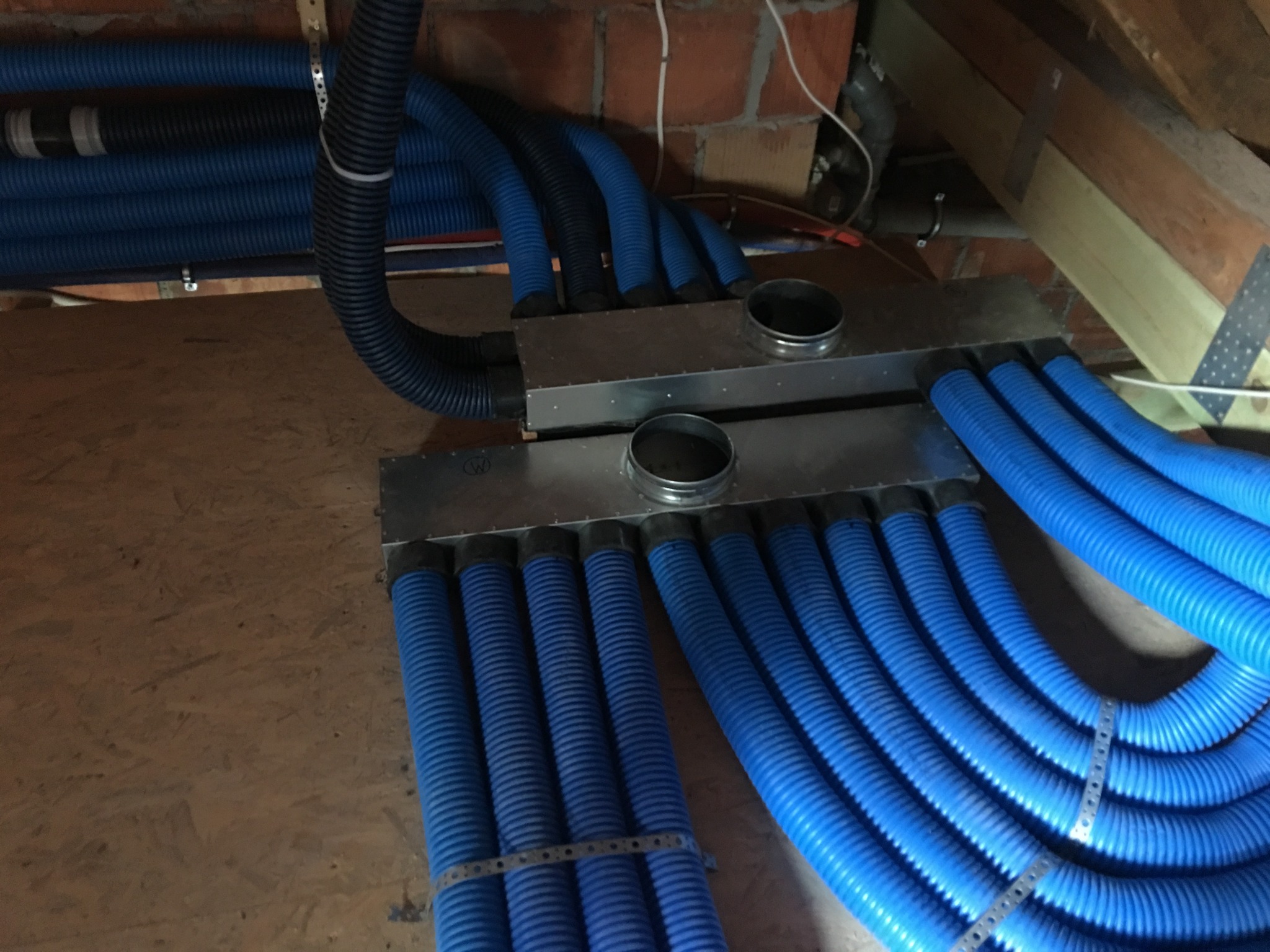 Rozdzielacz powietrza z licznymi niebieskimi przewodami wentylacyjnymi w surowym otoczeniu poddasza, widoczne elementy konstrukcji drewnianej i ceglanej, przewody spięte metalowymi obejmami.