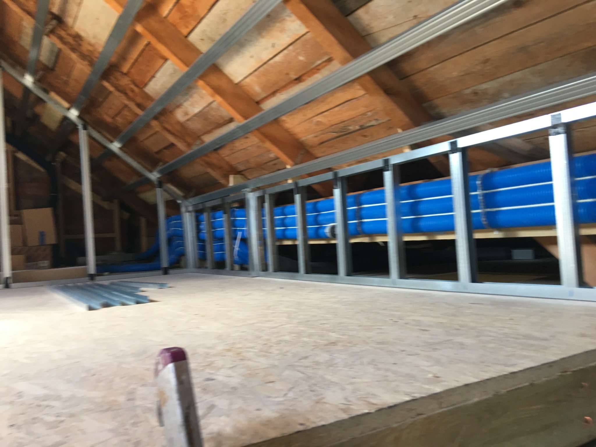 Poddasze z widoczną konstrukcją metalową i niebieskimi przewodami wentylacyjnymi, częściowo zamontowanymi pod drewnianym stropem, z perspektywy podłogi.