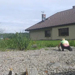 Kobieta w ogrodzie układa kamienne płyty na żwirowej powierzchni, w tle budynek mieszkalny i trawa ozdobna.