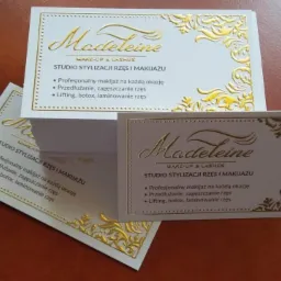 Stos wizytówek z tłoczonym złotym ornamentem i napisem 'Madeleine MAKE-UP & LASHES STUDIO STYLIZACJI RZĘS I MAKIJAŻU', prezentujących ofertę profesjonalnego makijażu, przedłużania i liftingu rzęs.