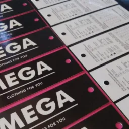 Partia czarno-białych etykiet odzieżowych z logo 'MEGA CLOTHING FOR YOU', część z instrukcjami prania, ułożonych rzędami na różowym podłożu.