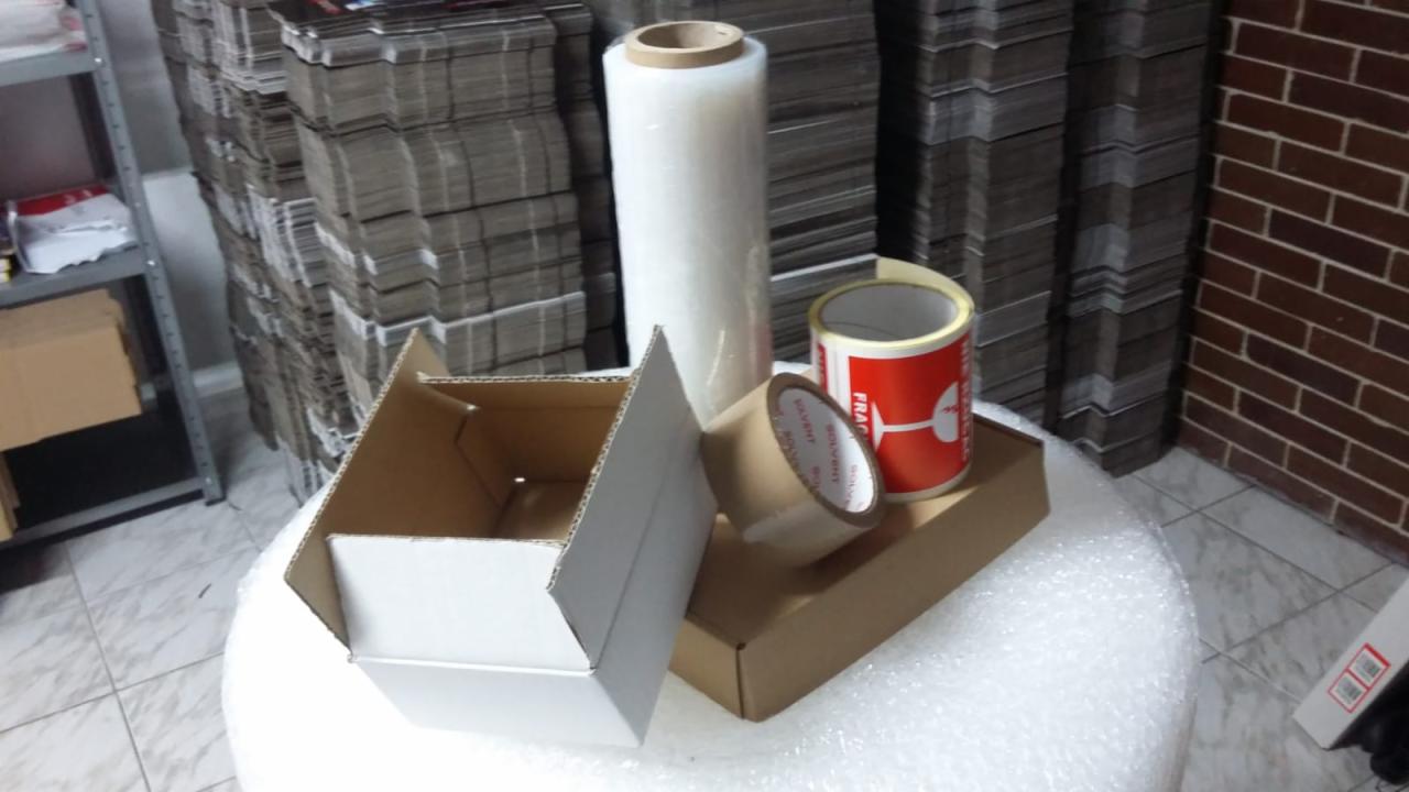 Rozłożone białe pudełko kartonowe, rolka folii stretch, taśma klejąca i rolka taśmy ostrzegawczej na stole z folii bąbelkowej, w tle stosy złożonych pudełek i ceglana ściana.