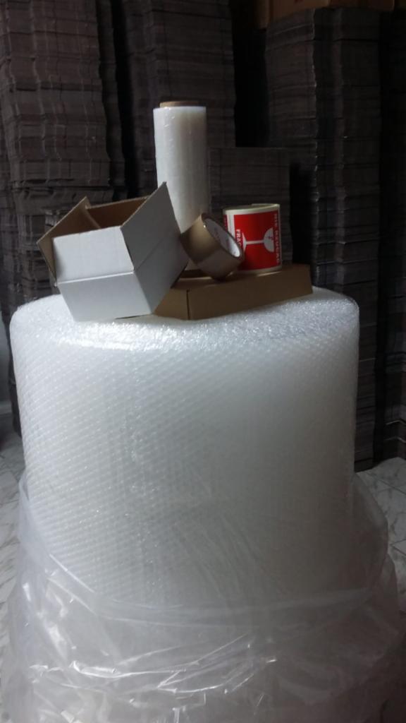 Stos kartonów w tle, na pierwszym planie rolka folii bąbelkowej, kartonowe pudełko otwarte, rolka taśmy klejącej i pudełko z oznaczeniem 'Fragile'.