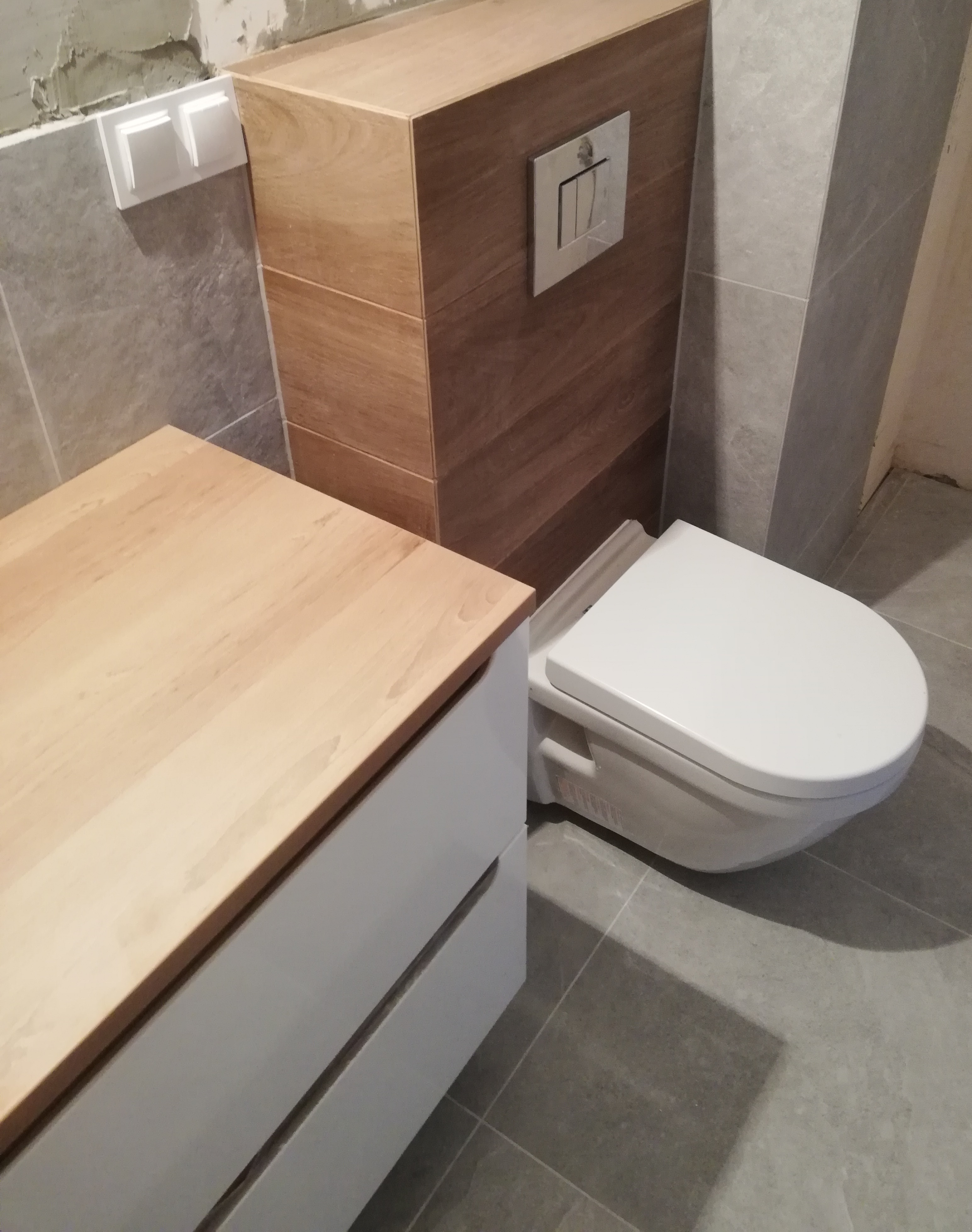 Nowoczesna toaleta podwieszana z drewnianą obudową stelaża i szarymi płytkami, obok szafka z jasnym drewnianym blatem, widoczny fragment niedokończonej ściany.