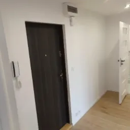 Wnętrze mieszkania po remoncie: widok na przedpokój z ciemnymi drzwiami wejściowymi, jasną podłogą i otwartymi drzwiami do łazienki z kabiną prysznicową.