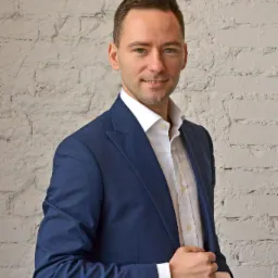 Portret uśmiechniętego agenta nieruchomości w granatowym garniturze na tle białej ściany z cegły.