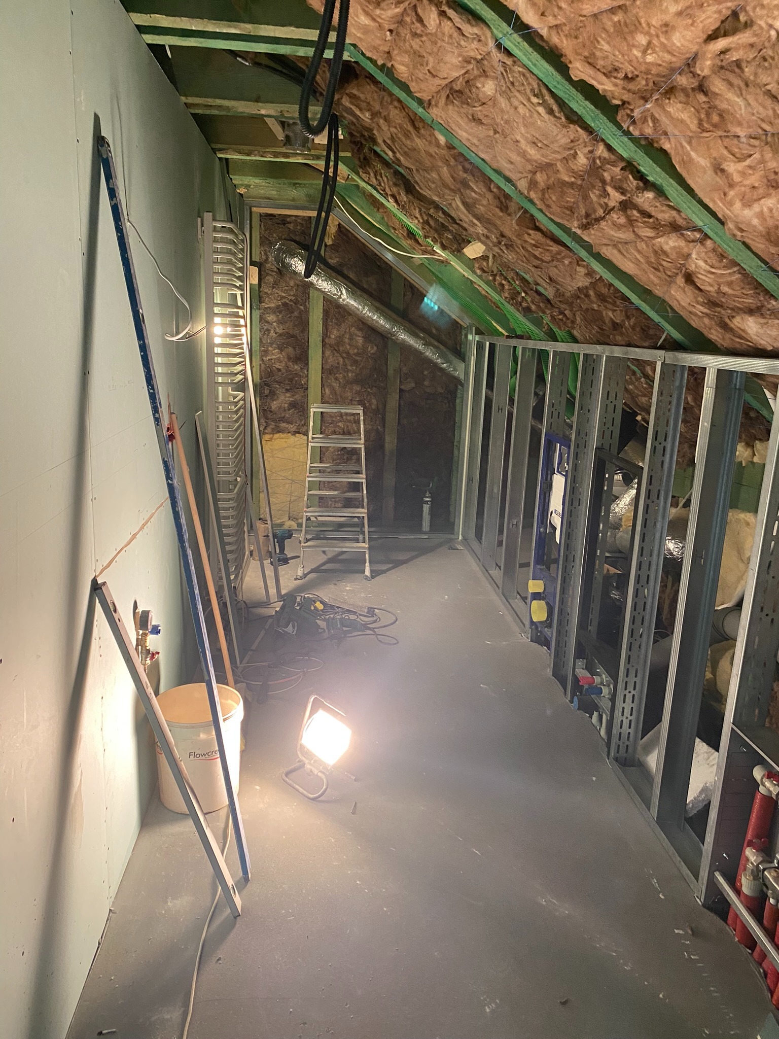 Poddasze w trakcie adaptacji: widoczne ocieplenie wełną mineralną, metalowa konstrukcja ścianki działowej, częściowo wykończona ściana z płyt gipsowo-kartonowych, narzędzia i drabina.